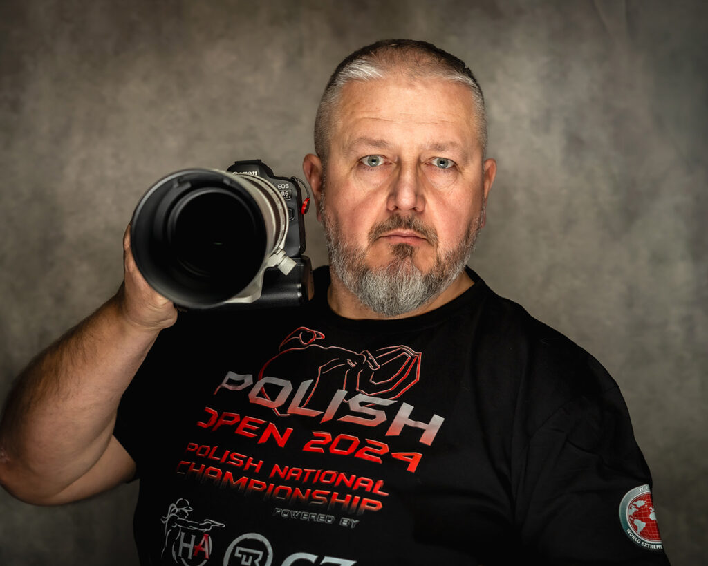 Portret fotografa w koszulce z zawodów IPSC, w ręku trzyma aparat z obiektywem
